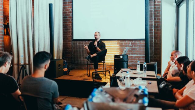 A conference with a man sitting on stage with a microphone and an audience listening.