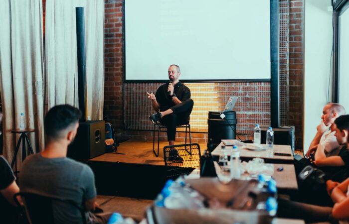 A conference with a man sitting on stage with a microphone and an audience listening.