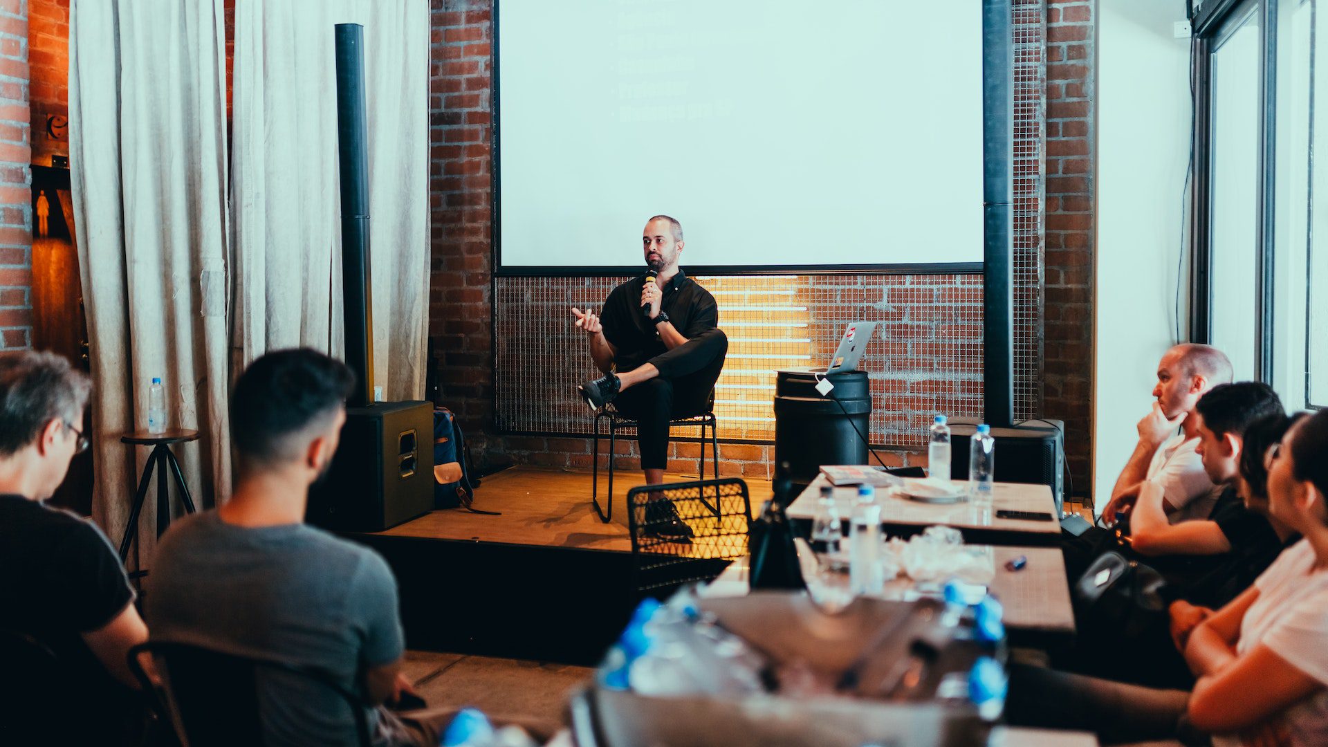 A conference with a man sitting on stage with a microphone and an audience listening.
