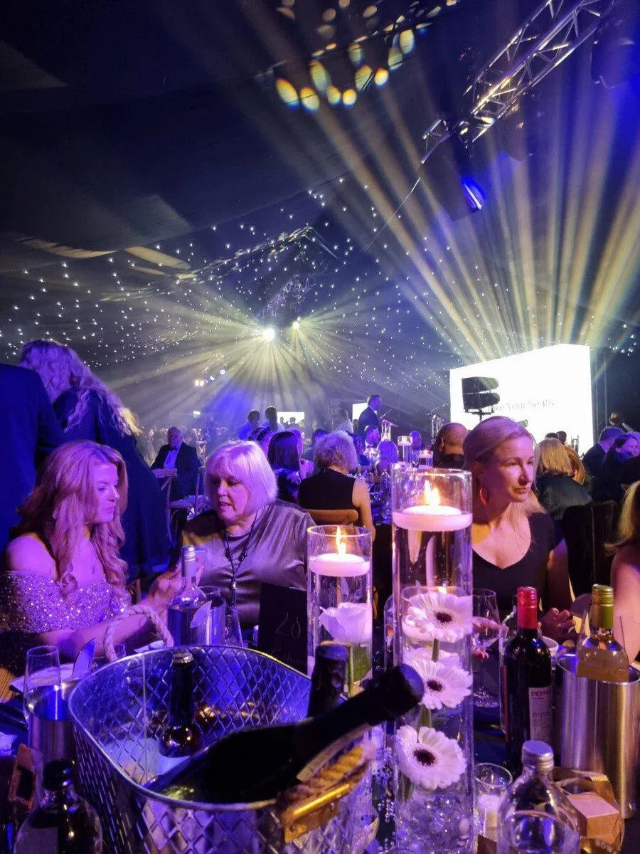 WrexhamComunityAndBusinessAward1 A group of people sit at a table, with dance lights in the background. In the foreground is a champagne flute and a champagne bucket