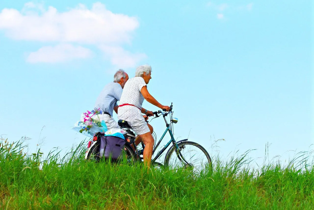 A two older people on bikes against a blue sky, with grass in the foreground
